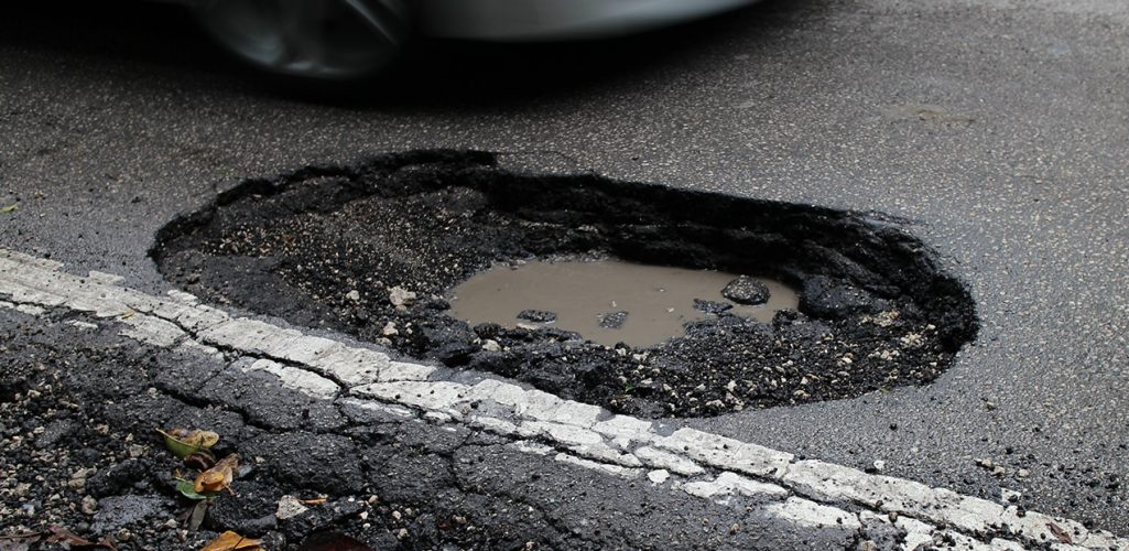 A very large pothole with a pool of dirty water inside it, deep in the surface of a road.