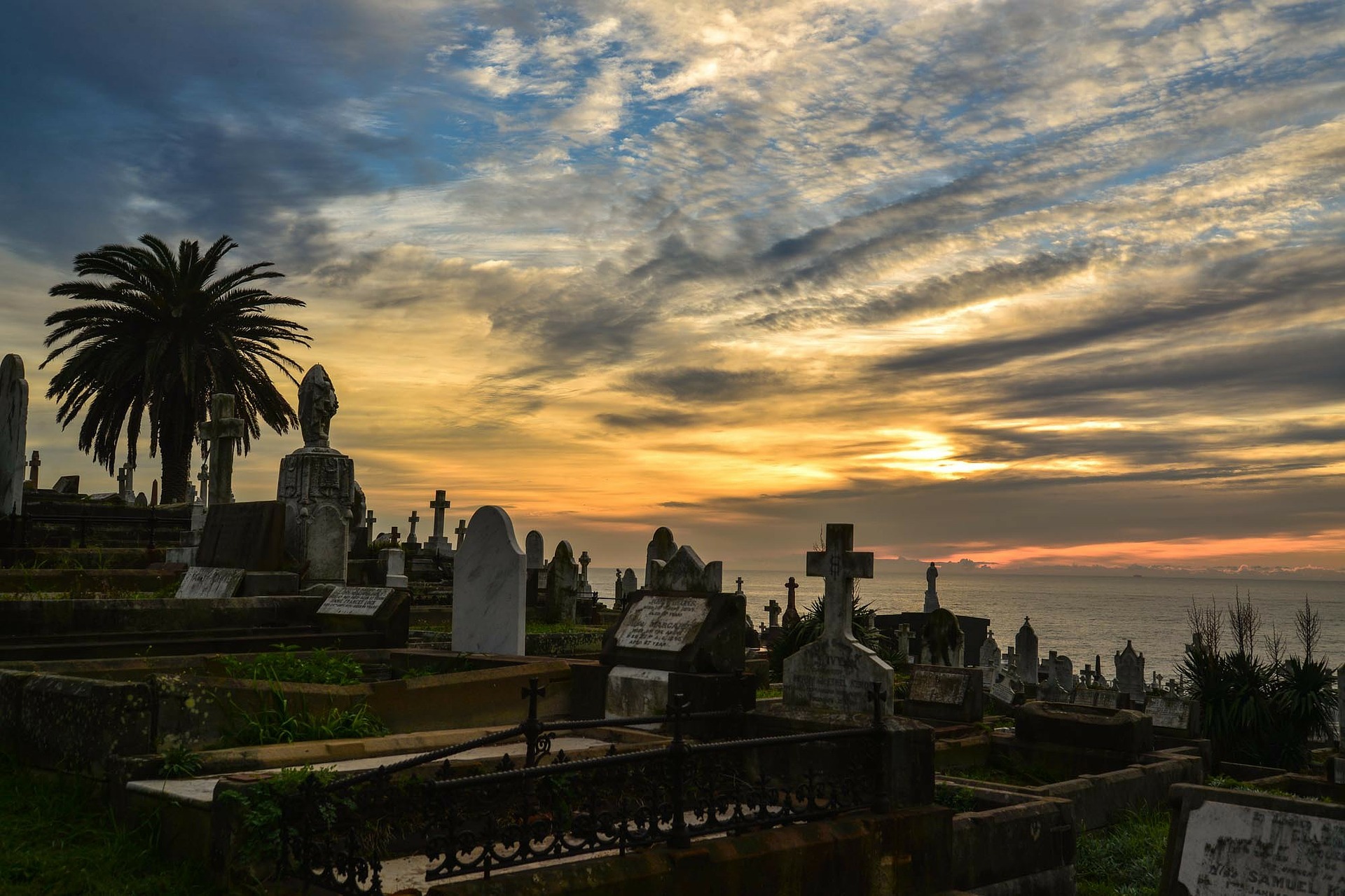 A graveyard with many various headstones stretches out in the foreground, beneath a hazy sunlit sky beside the sea.