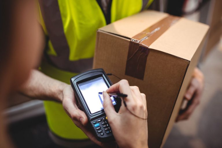 A courier wearing a high-visibility vest and holding a brown cardboard box and an e-signature reader awaits a signature from the recipient of the delivery.