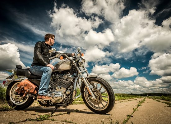 A high contrast photo of a man sat abreast a classic motorbike on an overgrown road beneath a cloudy blue sky.