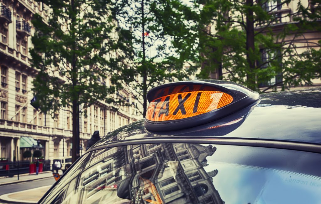 A black cab in a London street; the angle is looking at the orange taxi sign mounted on the roof, whilst trees and buildings are visible in the background.