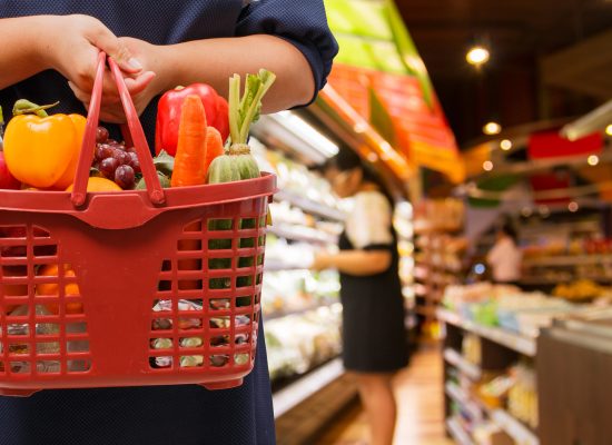 A shopper stands in the foreground of the picture holding a basket of vegetables and fruit, meanwhile in the background is an out of focus grocery store.