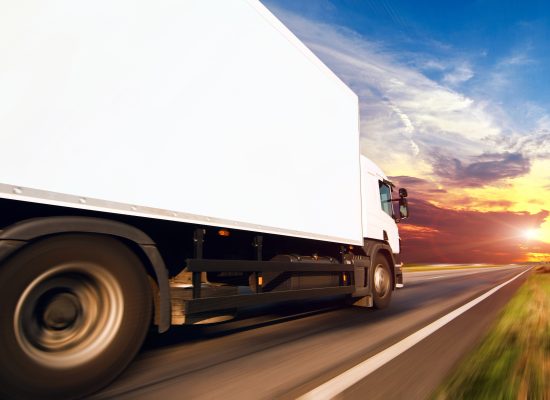 A white lorry driving along a straight flat road, beneath a beautiful sunset. The terrain is out of focus as a result of motion blur.