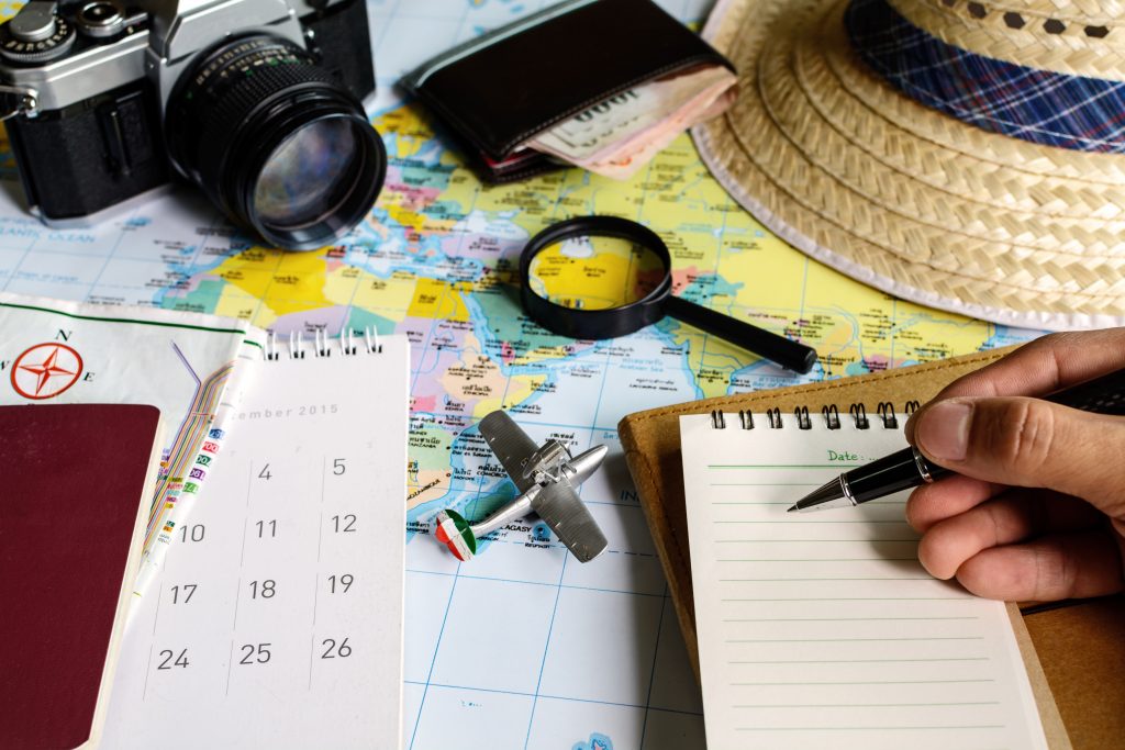 A collection of travel paraphernalia such as a map, straw hat. magnifying class, calendar, camera, wallet with money and a small aeroplane model. In the bottom right corner, a hand holds a pen, poised to write on a small notebook.