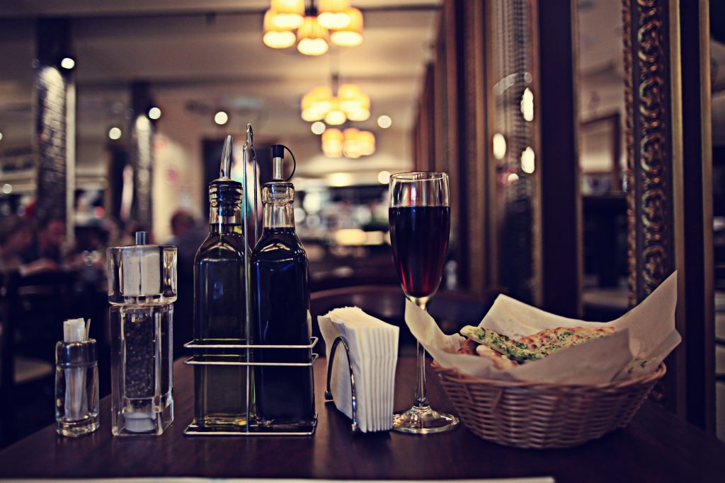 The foreground shows various table items at a restaurant. There is a sugar holder, salt and pepper, vinegar and olive oil, napkins, a glass of merlot wine and a small basket of bread.