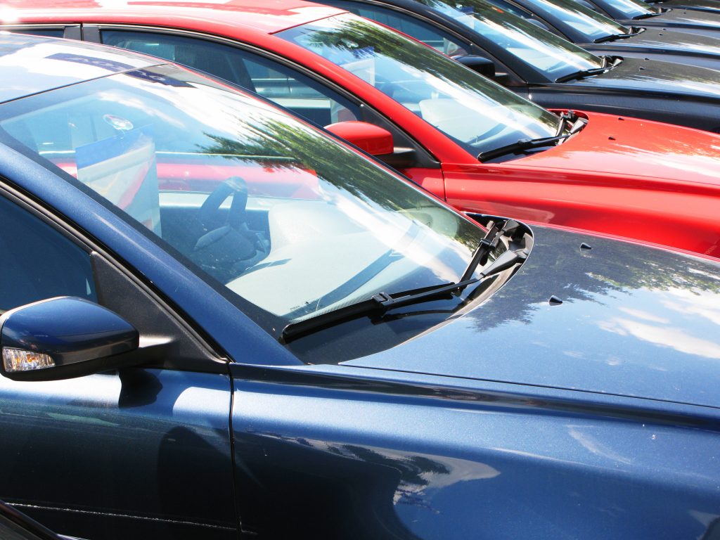 A row of blue and red cars on a carpark of forecourt; the camera angle is looking across the windscreens where you can see their is pricing information on display.
