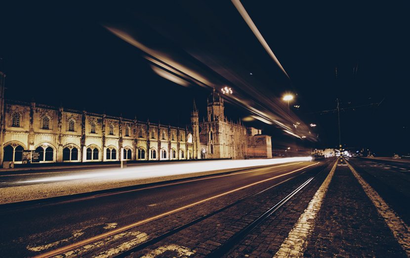 A sepia photo of a street at night time. There is a large gothic architectural building at the side of the road, and motion blurred traffic suggesting high speed vehicles.
