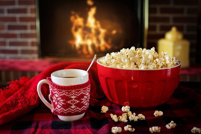 A bowl of popcorn, and a mug of coffee wearing a festive knitted cosy, sit in the foreground upon a tartan blanket, whilst a fireplace is out of focus blazing in the background,